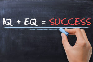 The image shows a hand writing on a chalkboard with the equation "IQ + EQ = SUCCESS." The letters "IQ" and "EQ" are written in white, while the word "SUCCESS" is written in bold red, emphasizing the idea that both intellectual intelligence (IQ) and emotional intelligence (EQ) contribute to achieving success.