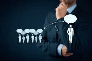 **Alt text**: A conceptual image illustrating leadership and influence. A man in a business suit, with his hand resting thoughtfully on his chin, stands in the background. In the foreground, a group of smaller figures in business attire are shown with thought bubbles above their heads. An arrow leads from their thought bubbles to a larger figure, representing a leader, who has a speech bubble above his head. This symbolizes the flow of ideas and communication from a group to a leader who speaks on their behalf. The image conveys the concept of leadership, decision-making, and influence in a corporate setting.