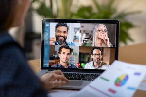 The image shows a woman participating in a video conference call on her laptop. The screen displays four colleagues in a virtual meeting, with two men, one woman, and a male host. The woman is holding a document with a pie chart in her hand, indicating that they are discussing work-related topics. The background suggests a home office or remote working environment.