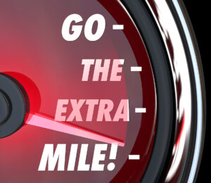 "Close-up of a speedometer-like gauge reading 'Go the Extra Mile!' in bold white letters, with the needle pointing to the highest level. The red background and sleek metallic edges emphasize motivation, commitment, and a push toward peak performance."