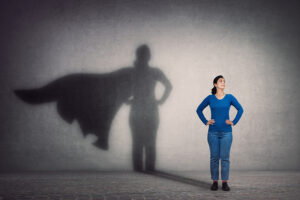 A confident woman stands with hands on her hips, facing forward. Behind her, a shadow on the wall reveals the silhouette of a superhero, symbolizing inner strength, potential, and the power to overcome challenges