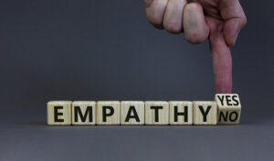 The image features wooden blocks spelling out the word "EMPATHY" in black letters. To the right, a hand points to a final block that is tilted, showing the words "YES" and "NO," suggesting a decision or choice regarding empathy. The background is a neutral gray, and the image emphasizes the importance of empathy as a key decision in personal or professional contexts.