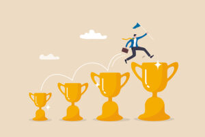 The image depicts a cartoon-style illustration of a person leaping from one gold trophy to a larger one in a progressive series of increasing-sized trophies. The individual, dressed in business attire, is holding a small flag and carrying a briefcase, symbolizing a journey toward success. Each leap between trophies represents steps in personal or professional achievement. The progression from smaller to larger trophies conveys the concept of building upon small wins to reach bigger goals and ultimate success. The overall tone of the image is motivational, emphasizing growth and continuous achievement.