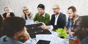 A diverse group of professionals engaged in a collaborative discussion around a table, examining a solar panel and various blueprints. The group is smiling and interacting, reflecting teamwork, innovation, and a focus on sustainable solutions in a modern workplace setting.
