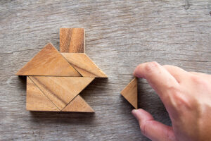 "Wooden tangram puzzle pieces forming a house shape on a rustic wooden surface, with a hand placing the final triangular piece to complete the structure."