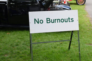 A white sign with green text reading "No Burnouts" is displayed on a grassy area, supported by a metal frame with chains securing it. In the background, classic cars, including a black vehicle with an open hood, are visible at what appears to be an outdoor car show or event