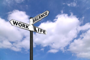 A signpost against a bright blue sky with clouds, featuring three directional signs labeled 'Work,' 'Life,' and 'Balance,' symbolizing the concept of work-life balance.